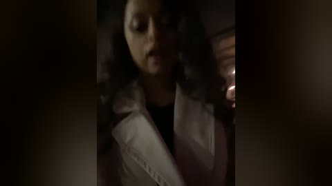 Media: A dimly lit video of a woman with curly black hair, wearing a white jacket over a black top, in a car, holding a lit cigarette.