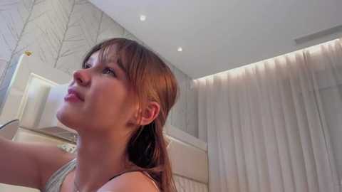 Media: Video of a young woman with fair skin, brown hair, and blue eyes, wearing a light-colored tank top, looking upwards in a modern, minimalist bathroom with white walls, a window with sheer curtains, and a leafless tree patterned wallpaper.