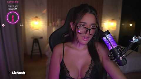 Media: Video of a slender, fair-skinned woman with long brown hair, wearing glasses and a black lace bra, sitting in a dark room with dim lighting, using a purple microphone.