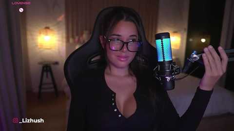 Media: A video of a woman with long dark hair, wearing glasses, black top, and a black hoodie, recording a video with a blue-lit microphone in a dimly lit bedroom with a bed and lamp in the background.