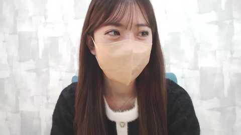 Media: Video of an Asian woman with long brown hair, wearing a white surgical mask and black cardigan, against a white and gray patterned backdrop.