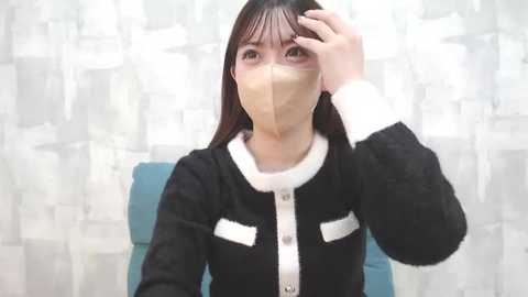 Media: Video of an East Asian woman with long black hair and light skin, wearing a black and white knit cardigan, beige mask, and touching her forehead. She sits on a teal chair against a white and grey patterned background.