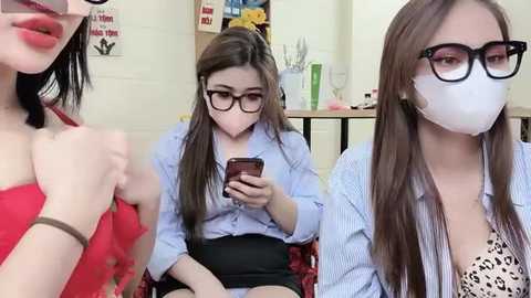 Media: Video of three young Asian women in a classroom, wearing masks, glasses, and casual attire; one texting, one reading, and one smiling.