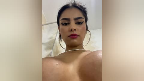 Media: Video of a woman with medium brown skin, dark hair, and large hoop earrings, wearing a pearl necklace, looking directly at the camera with a serious expression, in a shower.