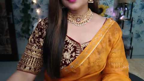 Media: Video of a woman in an orange sari with intricate gold embroidery, wearing a matching gold choker necklace. Background features a floral-patterned wall and a lamp.