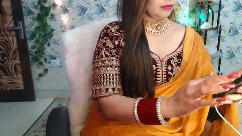 Media: Video of a woman in traditional Indian attire, including a maroon and gold embroidered blouse, an orange sari, and multiple gold jewelry pieces, seated on a white chair, holding a phone. The background features a floral-patterned wall and decorative objects.