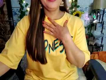 Media: Video of a woman with long brown hair, wearing a yellow T-shirt with \"PENELOPE\" in green letters, touching her chest, in a cozy room with floral decor and fairy lights.