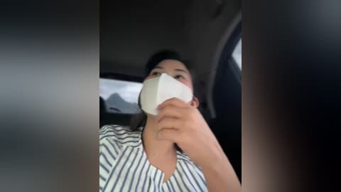 Media: A video of a woman wearing a blue and white striped shirt, with a white face mask, sitting in a car with blurred background.