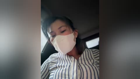 Media: A video of a woman wearing a white face mask, with dark hair tied back, in a car, dressed in a white and blue striped blouse.