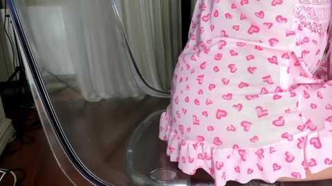 Media: Video of a person in a pink heart-patterned dress, standing in front of a see-through glass table. Background features a white curtain and wooden floor.