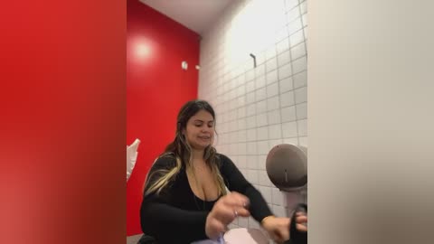 Media: Video of a plus-size woman with long, wavy hair, wearing a black top, sitting at a urinal in a public restroom with red and white tiled walls.