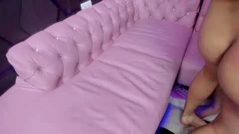 Media: Video of a nude person with a light skin tone, standing beside a pink, tufted leather couch with a diamond pattern. The background includes dark, textured furniture and a blue light source.