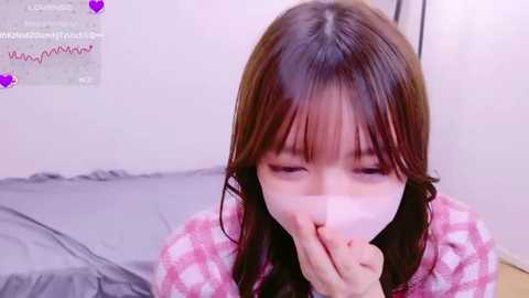 Media: A young Asian woman with long brown hair, wearing a pink checkered shirt, covering her mouth with her hand, indoors with a gray bed and white wall in the background.