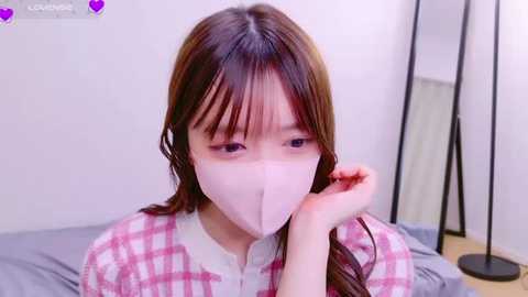 Media: Video of a young East Asian woman with long brown hair, wearing a pink plaid shirt and white face mask, sitting on a bed, in a simple bedroom with a black lamp and white wall.