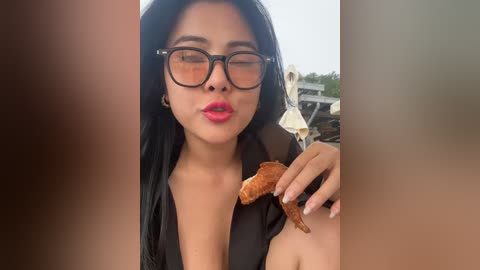 Media: Video of a young woman with long black hair, wearing oversized glasses, black top, and bright red lipstick, eating a chicken wing, with blurred background.