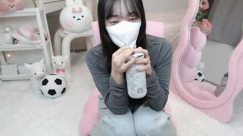 Media: Video of an East Asian woman with long black hair, wearing a gray sweater and a white face mask, sitting on a white carpet. She holds a baby bottle. The room is decorated with pastel pink and white decor, including plush toys and a mirror.