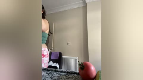 Media: A video of a woman with fair skin, wearing a green strapless top and pink patterned leggings, standing in a modern kitchen with beige walls, a white radiator, and a countertop with a tomato and a banana.