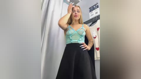 Media: Video of a blonde woman with fair skin, wearing a sheer mint-green lace camisole and black skirt, posing confidently in a fitting room with a white curtain and a red heart-patterned garment on a rack.