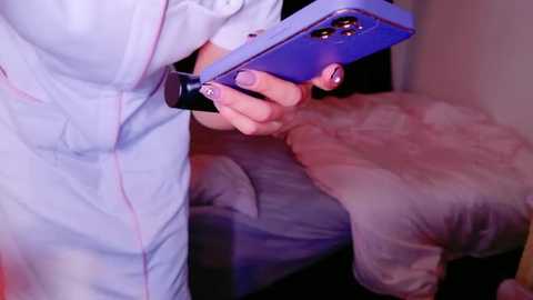 Media: Video of a person in a white medical gown holding a blue smartphone with a black case, sitting on a beige bed with a fluffy blanket.