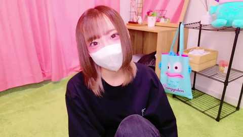 Media: Video of a young woman with shoulder-length light brown hair, wearing a black sweater and white face mask, sitting on a green carpeted floor. Background features pink curtains, a wooden desk, and a black metal shelf with toys and a blue tote bag.