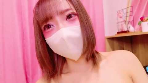 Media: Video of a young Asian woman with shoulder-length brown hair, wearing a white face mask, pink lipstick, and pink curtains in the background.