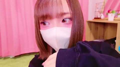 Media: Video of a young Asian woman with straight brown hair and bangs, wearing a white mask, dark top, and standing indoors against pink curtains and a wooden shelf with potted plants.