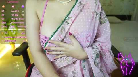 Media: Video of a light-skinned woman with a small breast size, wearing a pink floral robe with a green trim, exposing a pink bra. She is seated in a black chair, with a background featuring a dimly lit room and digital text on a screen.