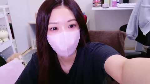 Media: Video of an East Asian woman with long dark hair, wearing a white mask, black top, and holding a selfie stick in a cluttered, brightly-lit room with white furniture and a stuffed animal.