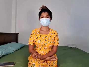 Media: Video of a young woman with medium skin tone, wearing a yellow floral dress, blue face mask, and brown hair tied up. She sits on a green bed in a simple, white-walled bedroom.