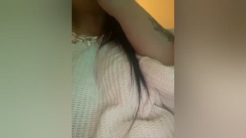 Media: Video of a person with fair skin and long, dark hair, wearing a white, textured sweater, lying on a bed, with a tattoo visible on their forearm. Background includes a yellow wall.