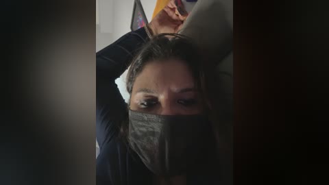 Media: Video of a woman in a dark room with her face obscured by a gray mask, revealing only her eyes, and her arm raised to her head.