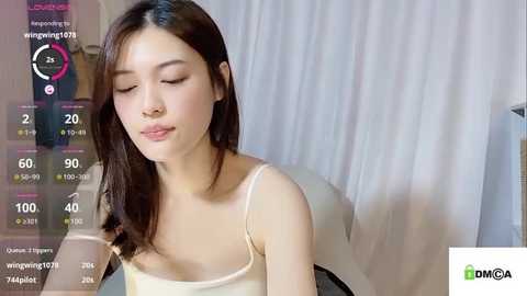 Media: A video of an Asian woman with long, straight dark hair, wearing a beige spaghetti-strap top, sitting indoors with a white curtain backdrop.