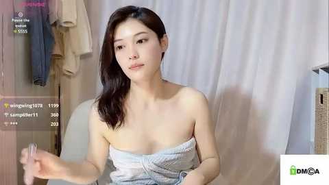 Media: Video of an East Asian woman with shoulder-length dark hair, wearing a strapless, light-colored dress, sitting in a room with hanging clothes and a white curtain background.