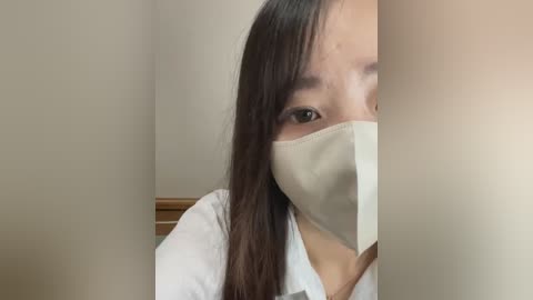 Media: A close-up video of a young Asian woman with long black hair, wearing a white surgical mask, peering from behind a beige curtain in a clinical setting.