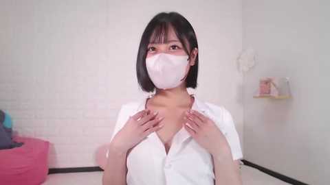 Media: Video of an Asian woman with short black hair, wearing a white face mask and a white button-up shirt, standing in a minimalist, white-walled room with a pink beanbag chair and shelves.