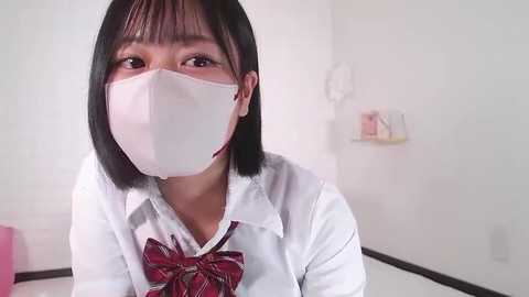 Media: Video of a young Asian woman with short black hair, wearing a white surgical mask, white blouse, and red plaid bow tie, standing in a white room with shelves in the background.