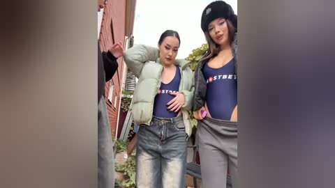 Media: Video of two Asian women in matching dark blue \"STRIPES\" one-piece swimsuits, light jackets, and jeans, posing outdoors near a red brick building.