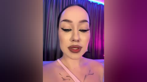 Media: Video of a young Asian woman with fair skin and long black hair, wearing black eyeliner, dark lipstick, and a light pink top, against a backdrop of dark curtains with purple and blue lights.