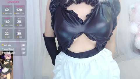 Media: Video of a woman wearing a revealing black maid outfit with a cut-out top, black gloves, and white apron, set against a soft-focus background.