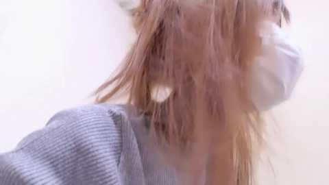 Media: Video of a person with shoulder-length, messy, light brown hair, wearing a gray sweater, and a white surgical mask. The background is blurred and neutral-colored, focusing on the person's face and hair.