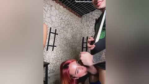 Media: A video of a young woman with red hair and tattoos, wearing a black shirt, playfully smacks a man's crotch in a dimly lit, stone-walled room with black metal furniture.