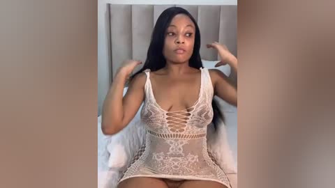 Media: A video of a Black woman with long black hair, wearing a revealing, white lace bodysuit, sitting on a bed with beige headboard, hands in her hair.