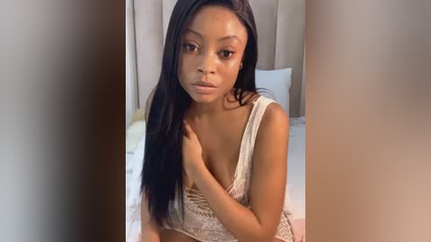 Media: Video of a young Black woman with smooth, medium-brown skin, long black hair, and a slender physique, wearing a revealing, beige lace top, sitting on a bed with white sheets in a softly lit, beige-tiled room.