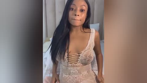 Media: Video of a young Black woman with long straight black hair, wearing a sheer lace dress with a plunging neckline, sitting on a bed in a beige room.