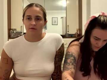 Media: Video of two women in a bathroom: one with blonde hair, white top, and tattoos, and another with dark hair, pink top, and tattoos. Background shows a mirror, towel rack, and framed art.