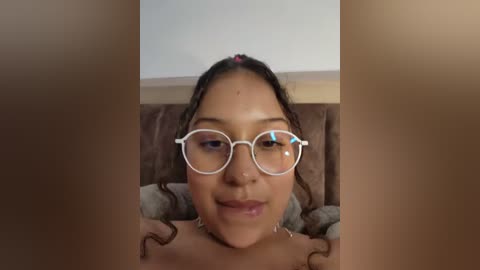 Media: A video of an Asian woman with medium-brown skin, wearing large glasses and a pink hair clip, smiling while lying on a bed with a brown blanket.