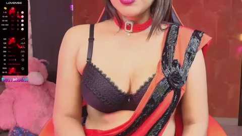 Media: Video of a South Asian woman with medium skin tone wearing a black lace bra, red sari, and red choker. Background includes a pink teddy bear and a \"Lovense\" screen.