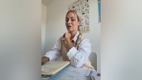 Media: Video of a young woman with light skin and blonde hair, wearing a white shirt and beige sweater, sitting at a desk with open books and papers.