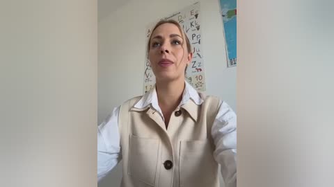 Media: Video of a blonde woman in a beige vest over a white shirt, standing in a brightly lit room with alphabet posters on the wall.