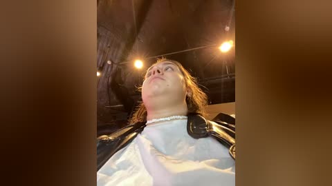 Media: Video of a person with shoulder-length light brown hair, wearing a white shirt and black leather jacket, standing in a dimly lit, cave-like setting with string lights.